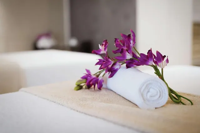 A rolled white towel and purple orchid stems rest on a massage table in a spa treatment room.