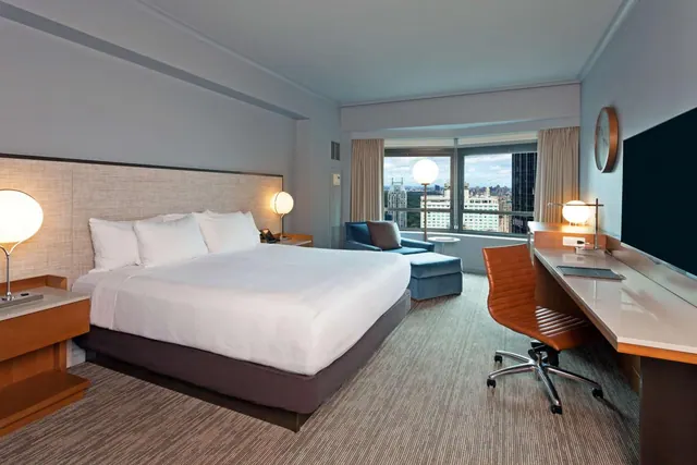 A hotel bedroom with a king bed, a blue armchair with an ottoman, and a wall-mounted television beside a desk, with a window showing a city view.