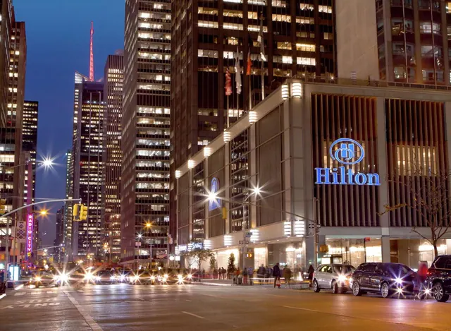 Night view of a Hilton hotel building on a city street with illuminated signage, parked cars, and a few pedestrians.