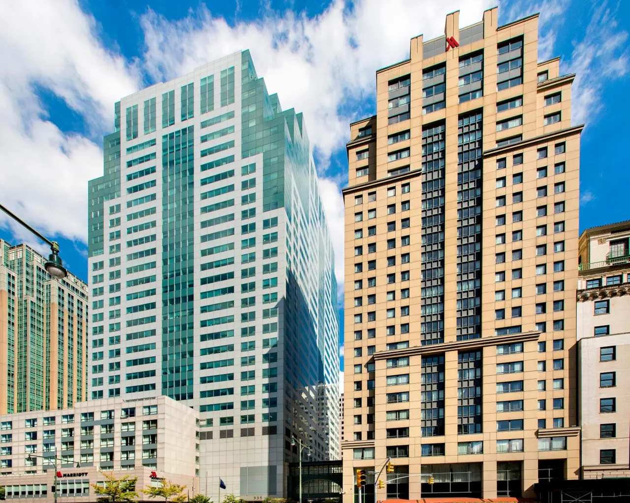Exterior view of two tall towers, one with glass cladding and one with tan stone facade, with a Marriott sign at street level under a blue sky with clouds.