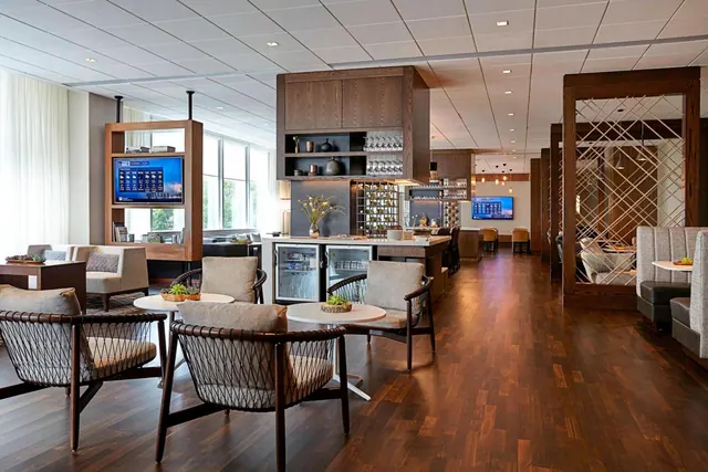 A lounge with upholstered armchairs and round tables on a wooden floor, with a bar counter holding glassware and refrigerators, and wall-mounted televisions.