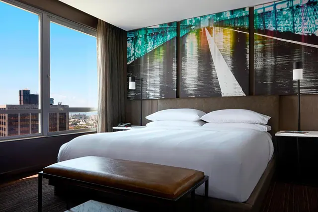 A king bed with white linens sits in front of a large window showing city buildings, with a triptych of a bridge artwork on the wall above the headboard.