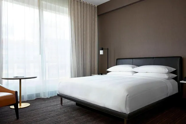 A made king bed with white linen sits beside a sheer-curtained window, with a round side table and a leather armchair at the left edge.