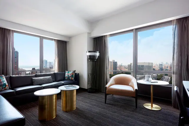 A living area with a black leather sofa, a tan armchair, gold-based side tables and floor-to-ceiling windows overlooking city buildings and a river.