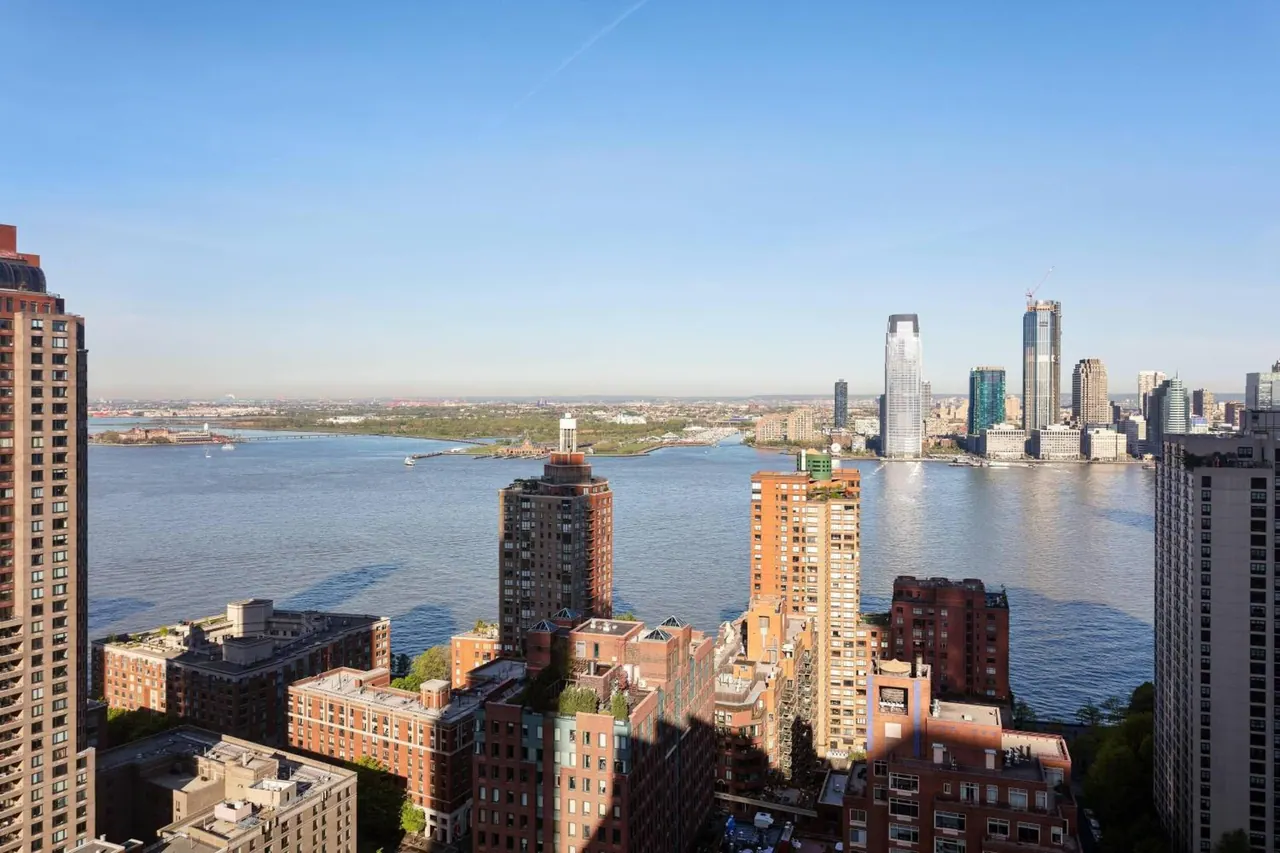 View from a high floor over the Hudson River toward a city skyline, with residential towers and brick buildings in the foreground.