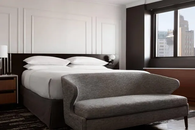 A bedroom with a made king bed, an upholstered bench at its foot, a wood credenza, and a window showing city buildings.