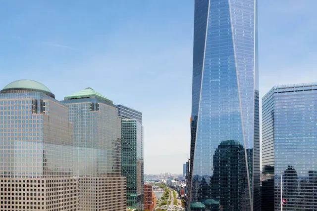 View of One World Trade Center and neighbouring glass office towers under a clear blue sky, with a street and waterway visible in the distance.