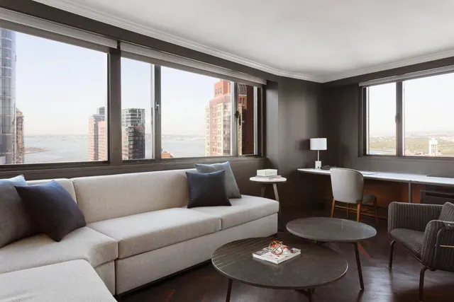 A living area with a light sectional sofa, dark round coffee tables, a desk by the window, and large windows showing city towers and water.