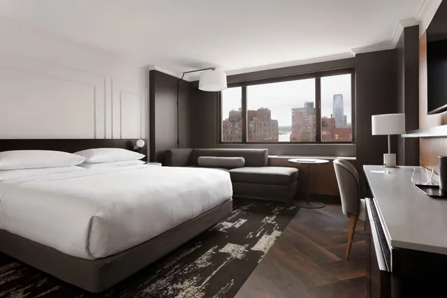 A king bed with white linens sits beside a grey sofa and a desk with a chair, and a window beyond shows brick and glass city buildings.