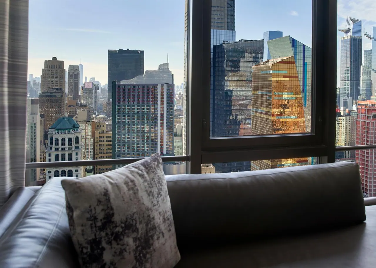 A leather sofa with a patterned cushion sits in front of a large window overlooking high-rise buildings in the city.