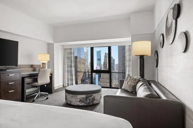 A hotel room with a large bed in the foreground, a grey sofa, a round ottoman, a desk with a chair and a television, and a window overlooking city skyscrapers.