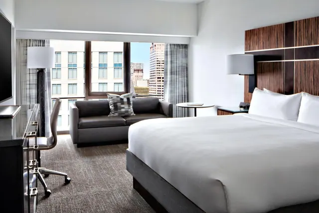 A hotel bedroom with a king bed, a grey sofa, a work desk and chair, and a large window looking out over city buildings.