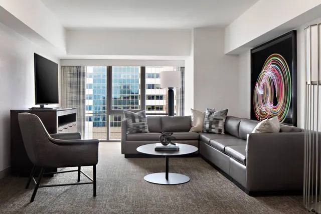 A staged living room with a grey sectional sofa, an armchair, a wall-mounted television and a large window showing city high-rises.