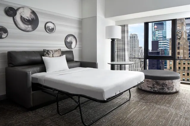A grey sofa bed is pulled out into a made bed in a room with floor-to-ceiling windows overlooking city buildings.