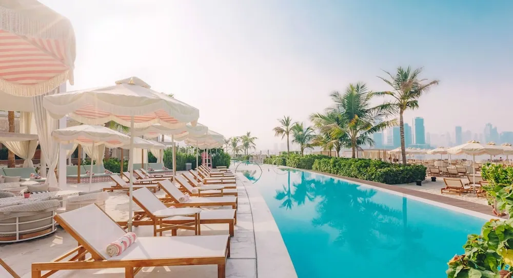 A large swimming pool lined with white sun loungers and striped umbrellas, with palm trees and a hazy city skyline in the background.