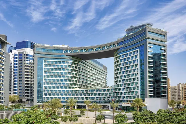 Exterior of a modern hotel building with a curved glass and patterned facade, a cantilevered bridge linking two sections, palm trees in front, and a blue sky with thin clouds.