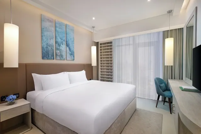 A bedroom with a made white king bed, a teal armchair, a wall-mounted television and a desk, with sheer-curtained floor-to-ceiling windows.