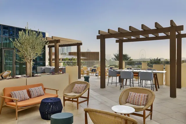 A furnished rooftop terrace with wicker chairs, an orange sofa, high tables under wooden pergolas, and a view of city towers and the Ain Dubai ferris wheel.