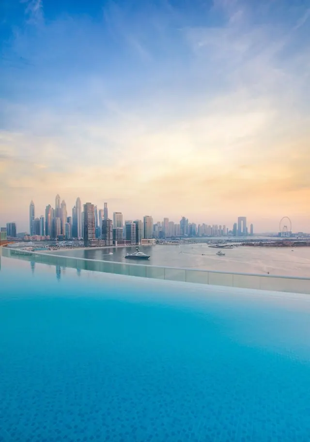 An infinity pool overlooks a city skyline across the water, with a large observation wheel visible on the right at dusk.