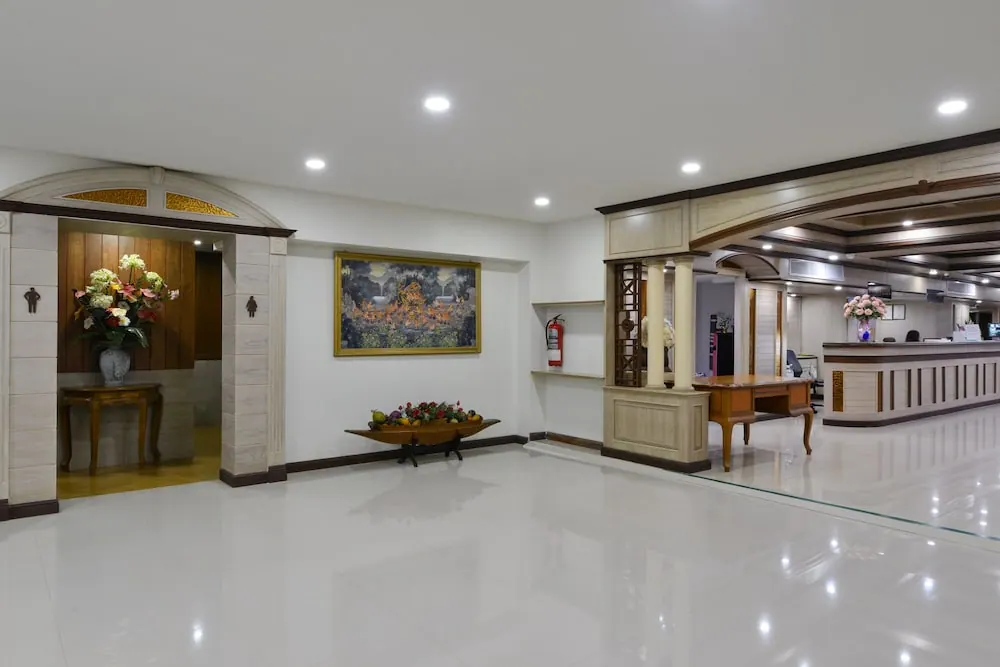 A hotel lobby with a reception desk on the right, a framed painting on the wall, a flower arrangement in an archway at left, and a low wooden table with a fruit centrepiece on a glossy white floor.