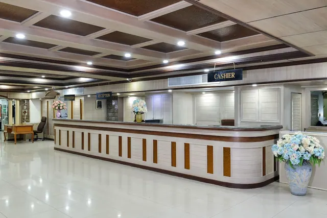 A long curved reception counter with RECEPTION and CASHIER signs sits under a coffered wooden ceiling, with flower arrangements on the polished tile floor and a wooden desk on the left.