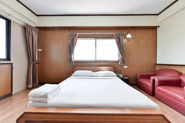 A bedroom with a double bed with white linens and folded towels, red leather armchairs, and wood-panelled walls around a window.
