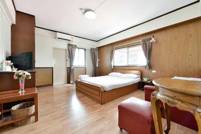 A double bed with white linen sits against a wood-panelled wall beside a window, with a red ottoman and a round wooden table in the foreground.