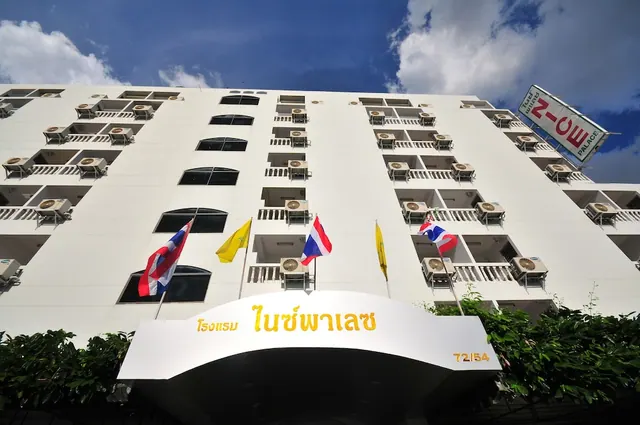Low-angle view of a white multi-storey hotel building with balconies and air conditioning units, Thai and royal yellow flags, and a Thai-language entrance canopy sign.