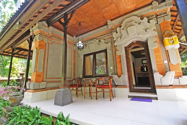 A covered terrace with carved stone and orange-painted Balinese detailing, wooden chairs with red cushions, a small table, and a carved doorway leading into a room.