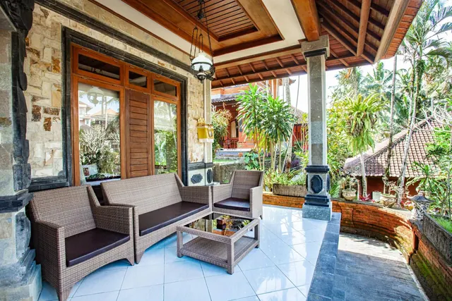 A covered veranda with wicker sofas, an armchair and a glass coffee table, with stone walls, timber doors and tropical garden beyond.