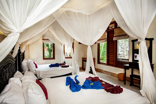 A bedroom with a canopy bed draped in white mosquito netting, a second bed in the background, towel animals on the bed, and windows looking out onto greenery.