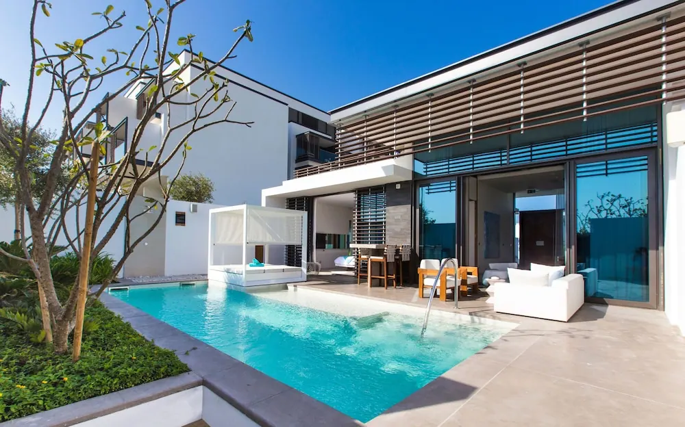 A private swimming pool sits beside a covered daybed and a white outdoor sofa on a patio in front of a villa with glass sliding doors.