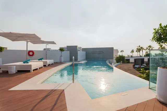A rooftop swimming pool with white sun loungers, teal towels and shade umbrellas on a wooden deck.