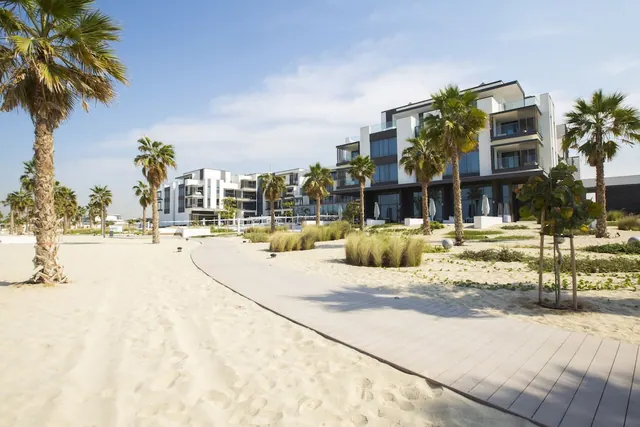 A wooden boardwalk crosses sand between palm trees and a white multi-storey building with balconies.