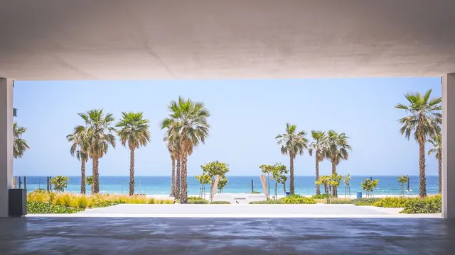 A covered concrete walkway frames a view of palm trees, a sandy beach and the sea under a clear sky.