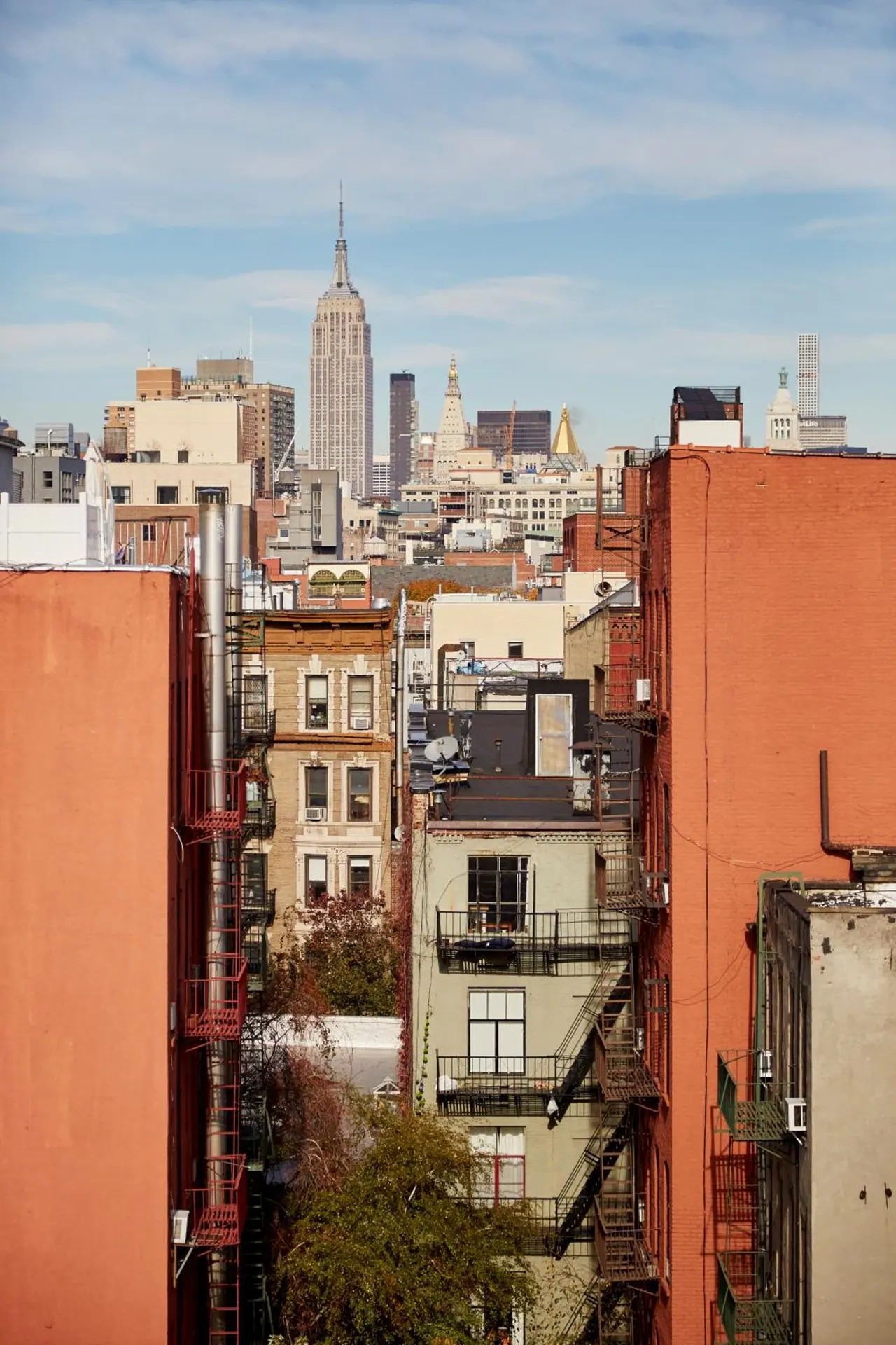 A view down a narrow street between brick buildings with fire escapes, with the Empire State Building visible in the distance under a partly cloudy sky.