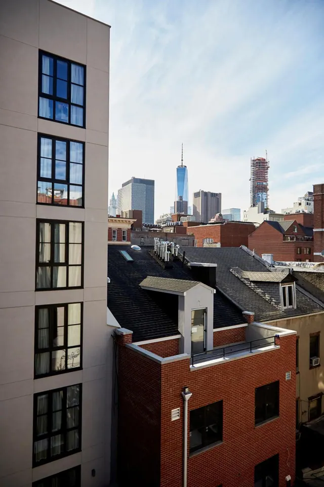 View over neighbouring rooftops and brick buildings toward the Lower Manhattan skyline, with One World Trade Center visible in the distance under a partly cloudy sky.