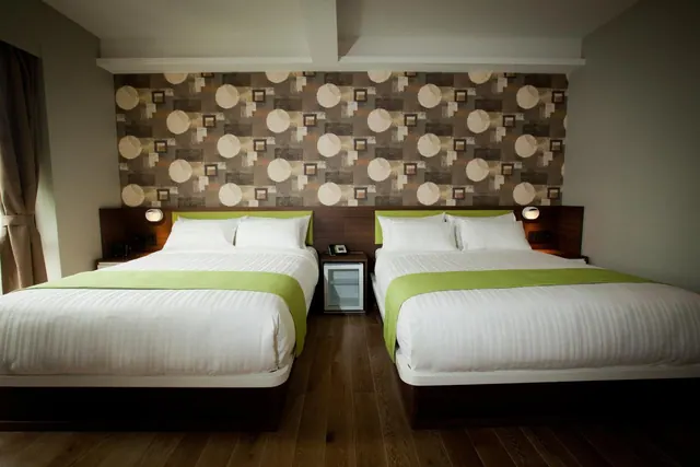 Two double beds with white linen and green runners stand in front of a patterned feature wall, with a small fridge between them.