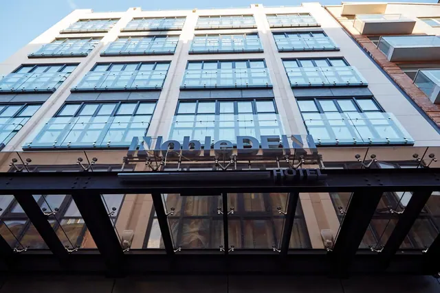 Low-angle view of the glass-fronted facade of the NobleDEN Hotel building, with its name sign above the entrance canopy.