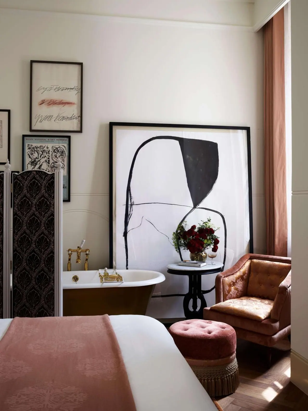A bedroom with a freestanding bathtub beside a bed, a large abstract artwork on the wall, and a pink armchair with a velvet ottoman.