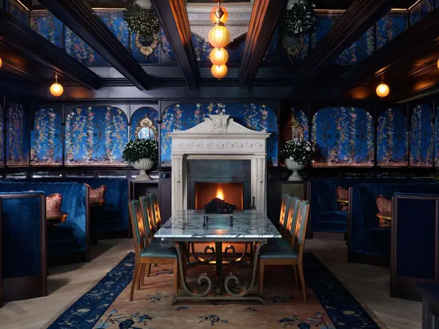 A dimly lit dining room with blue floral wallpaper, velvet banquettes, a marble-topped table and a white stone fireplace.