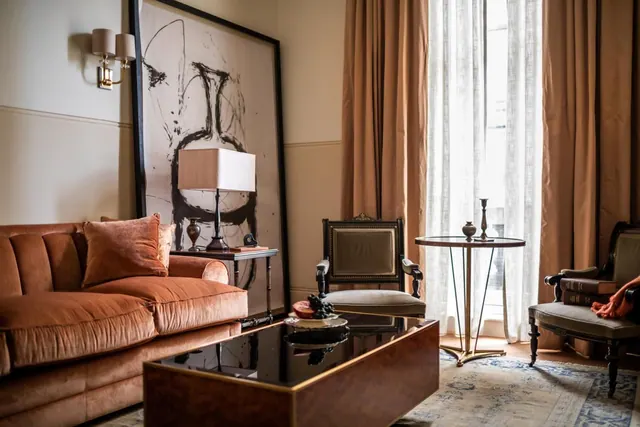 A living area with an orange velvet sofa, a large framed abstract artwork, a table lamp, a black glass coffee table and two upholstered chairs beside a window with sheer curtains.