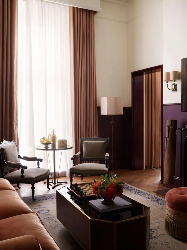 A living area with a velvet sofa, two armchairs, a small round table by tall curtained windows, a floor lamp and a coffee table with a chess set and flowers.