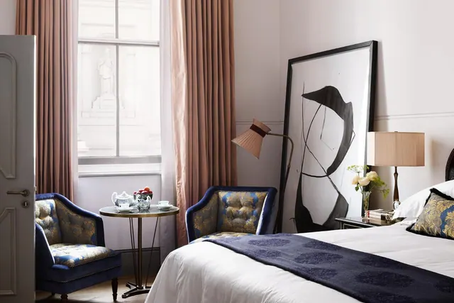 A bedroom with a made double bed, two blue armchairs, a round table with a teapot and cups, a tall window with pink curtains, and a large framed abstract artwork.