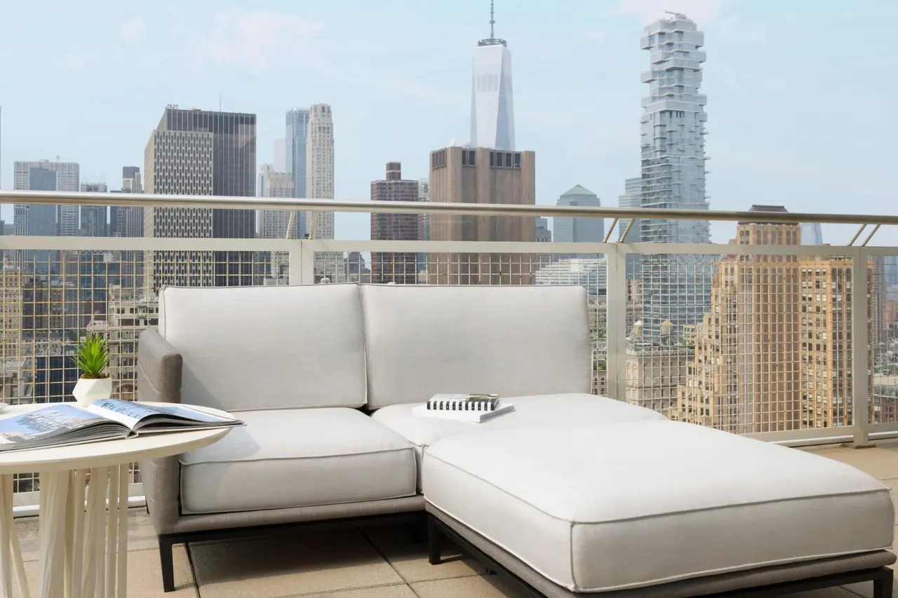 A terrace with a light grey sectional sofa, a side table with a magazine and a small potted plant, and a view of the Manhattan skyline including One World Trade Center.