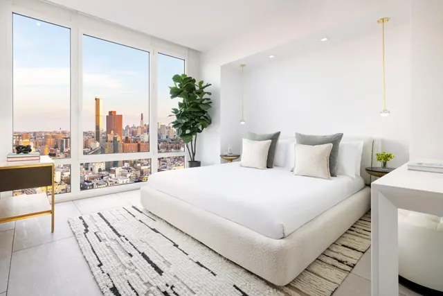 A bedroom with a white upholstered king bed, floor-to-ceiling windows overlooking a city skyline, a white desk and a potted fiddle-leaf fig plant.