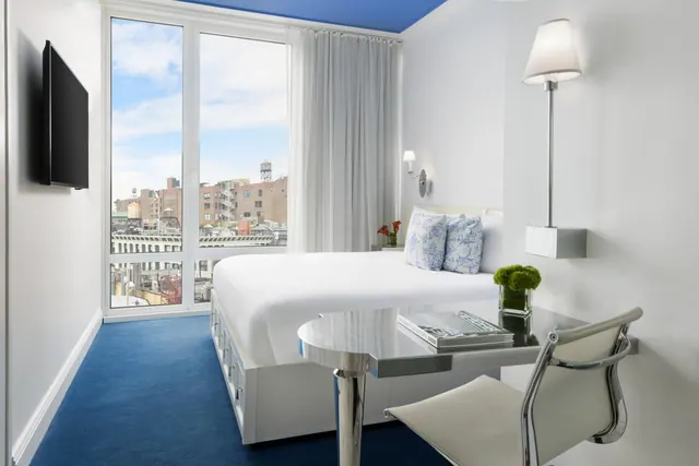A bedroom with a made king bed, a glass desk with a white chair, a wall-mounted television, and floor-to-ceiling windows looking out over city buildings and a water tower.