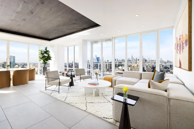 A living room with a beige sectional sofa, white coffee table and floor-to-ceiling windows looking out over a city skyline that includes the Empire State Building.