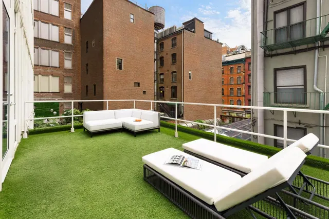 A furnished rooftop terrace with artificial turf, a white sectional sofa, two sun loungers and a magazine, with brick buildings beyond the railing.