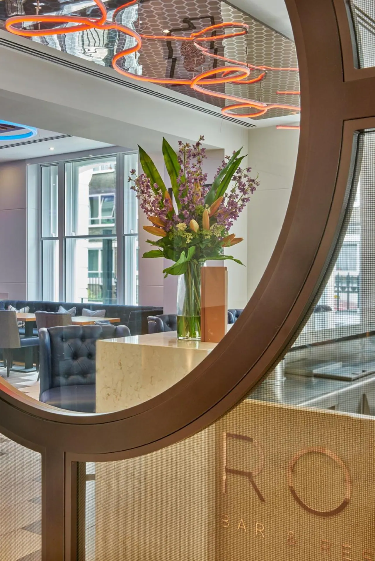 A circular bronze-framed screen in the foreground frames a bar and restaurant seating area with tufted chairs, a flower arrangement on a stone counter, and neon lighting on the ceiling.